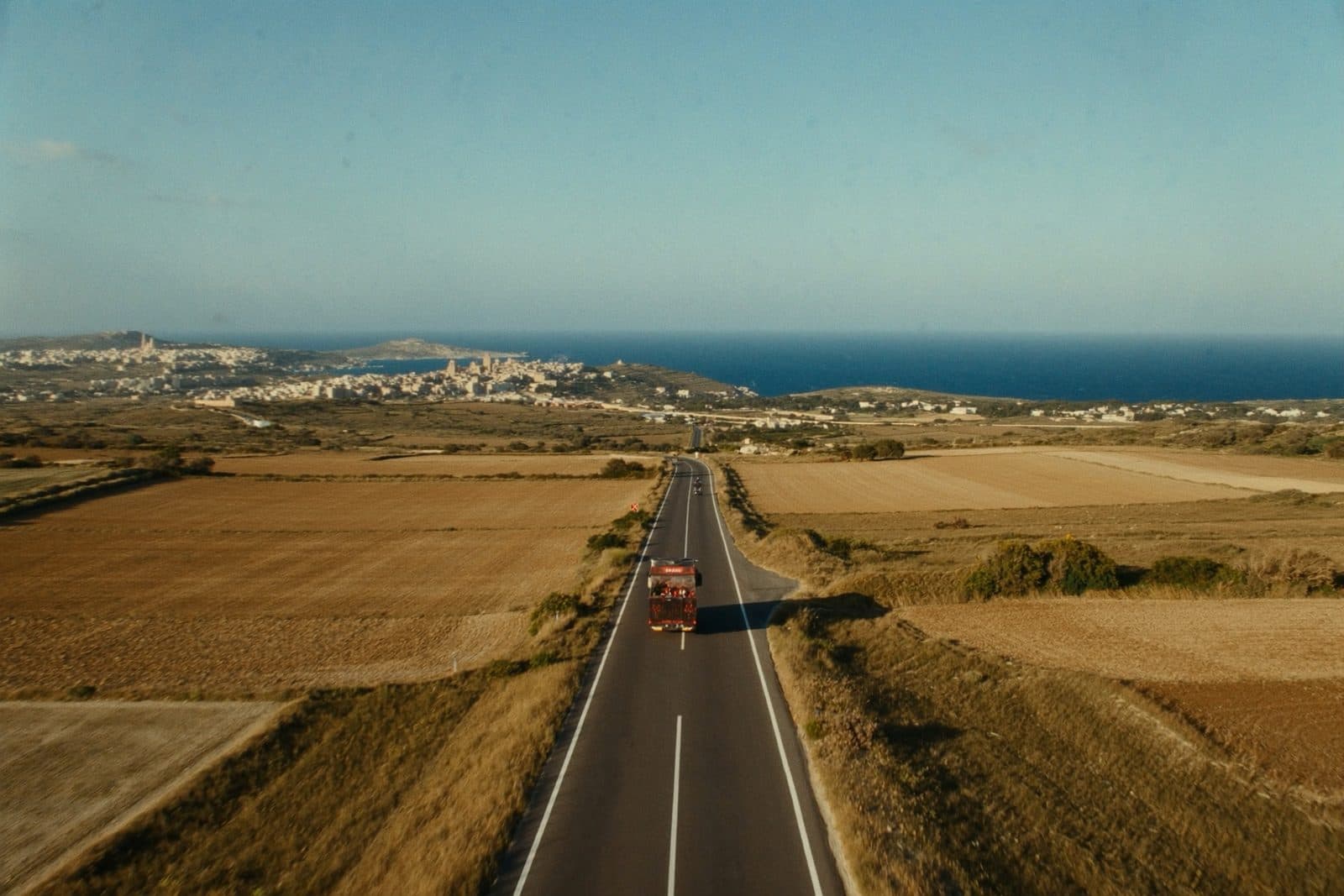 A sunlit road leading from the Maltese countryside toward a limestone town and the sea